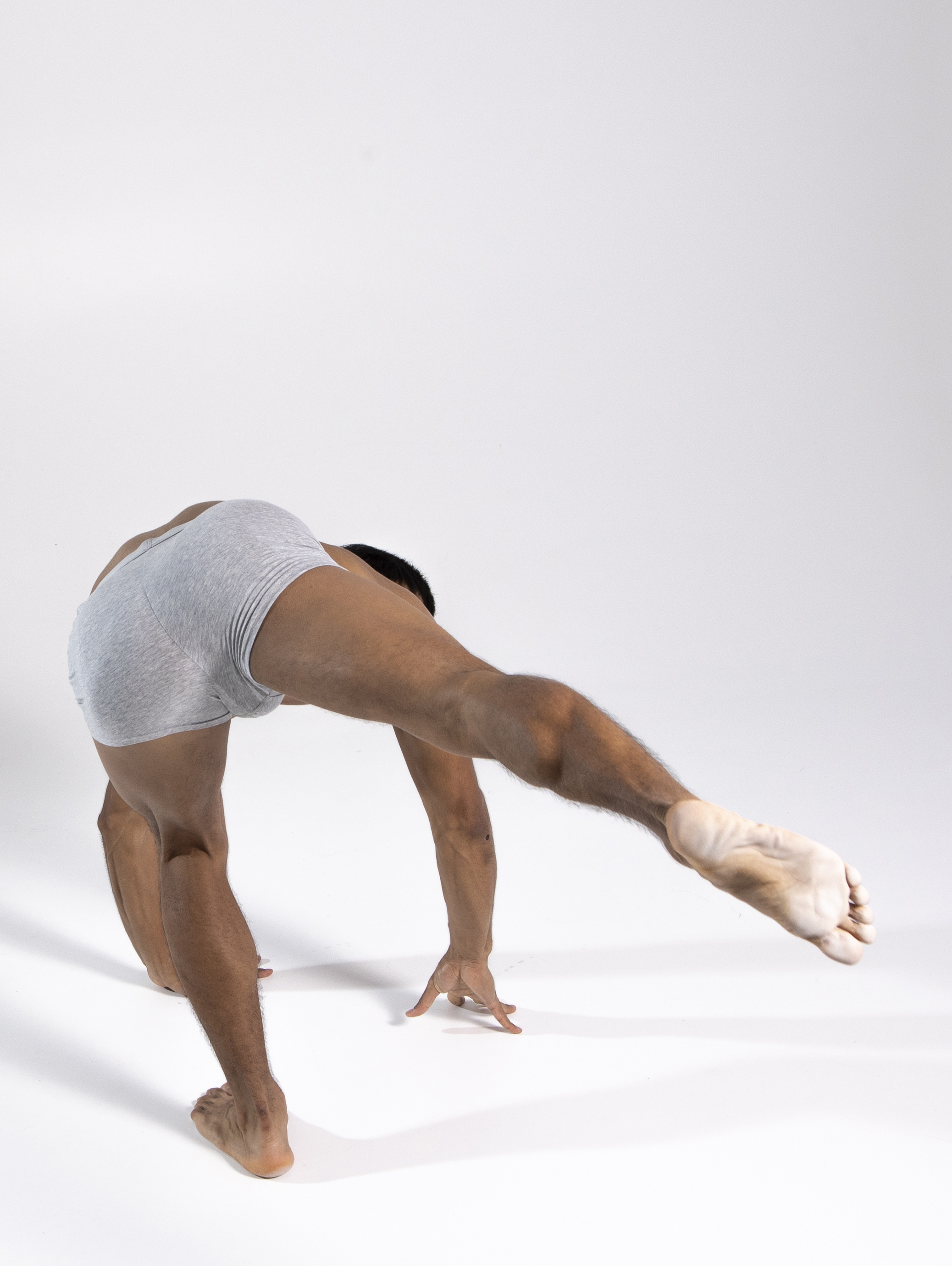 a man in gray underwear is posed in a studio shot against a white background. he is barefoot and bent over backwards, with one leg extended straight behind him, and his hands on the floor. the man is muscular and athletic looking. he has a skin condition, vitiligo, on his foot. he appears to be a dancer or acrobat, showing great flexibility.