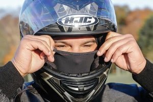 a closeup portrait of a man wearing a motorcycle helmet and a face mask. he is looking into the camera, with a serious expression on his face. the helmet is a black fullface helmet, with a clear visor. the face mask is a black balaclava. the man is wearing a black jacket and gloves. the background is out of focus.