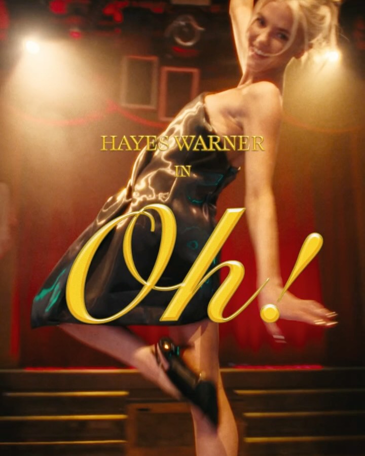 image shows a promotional poster for hayes warner in "oh!". a fairskinned woman with blonde hair in a black dress strikes a dynamic pose on stage with an excited expression. the text "hayes warner in oh!" is prominently displayed over the woman. spotlights shine down on the stage.