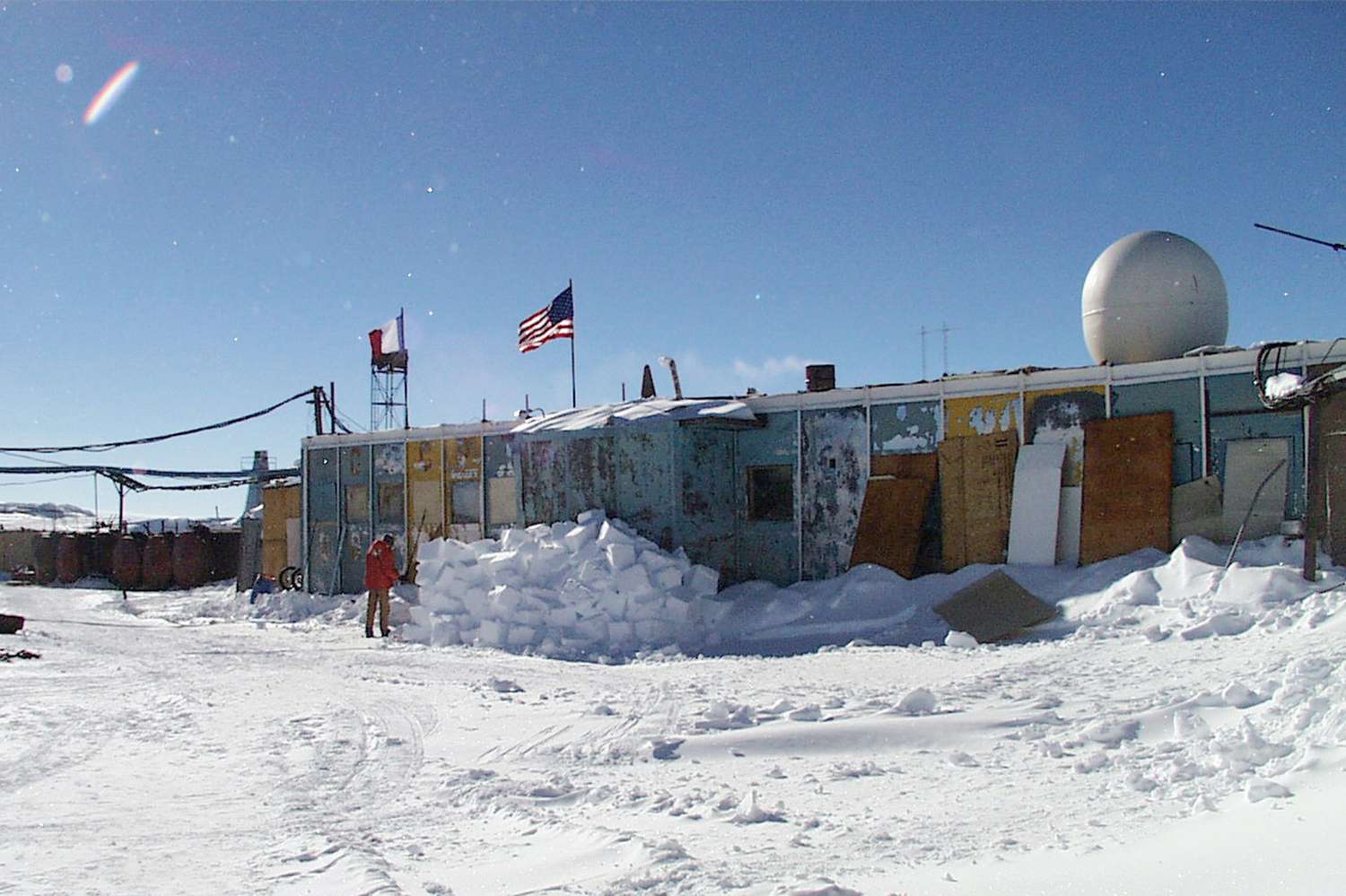 a view of a research station in antarctica, with snow covering the ground and buildings. the american flag is flying in the wind, and a snowcovered mountain range is visible in the background. the sky is blue and clear, with no clouds. this is a typical scene of the harsh conditions that researchers face in antarctica. the snow is so deep that it covers everything, and the air is so cold that it is difficult to breathe. but despite the challenges, researchers continue to work in antarctica to learn more about the planet and its climate.