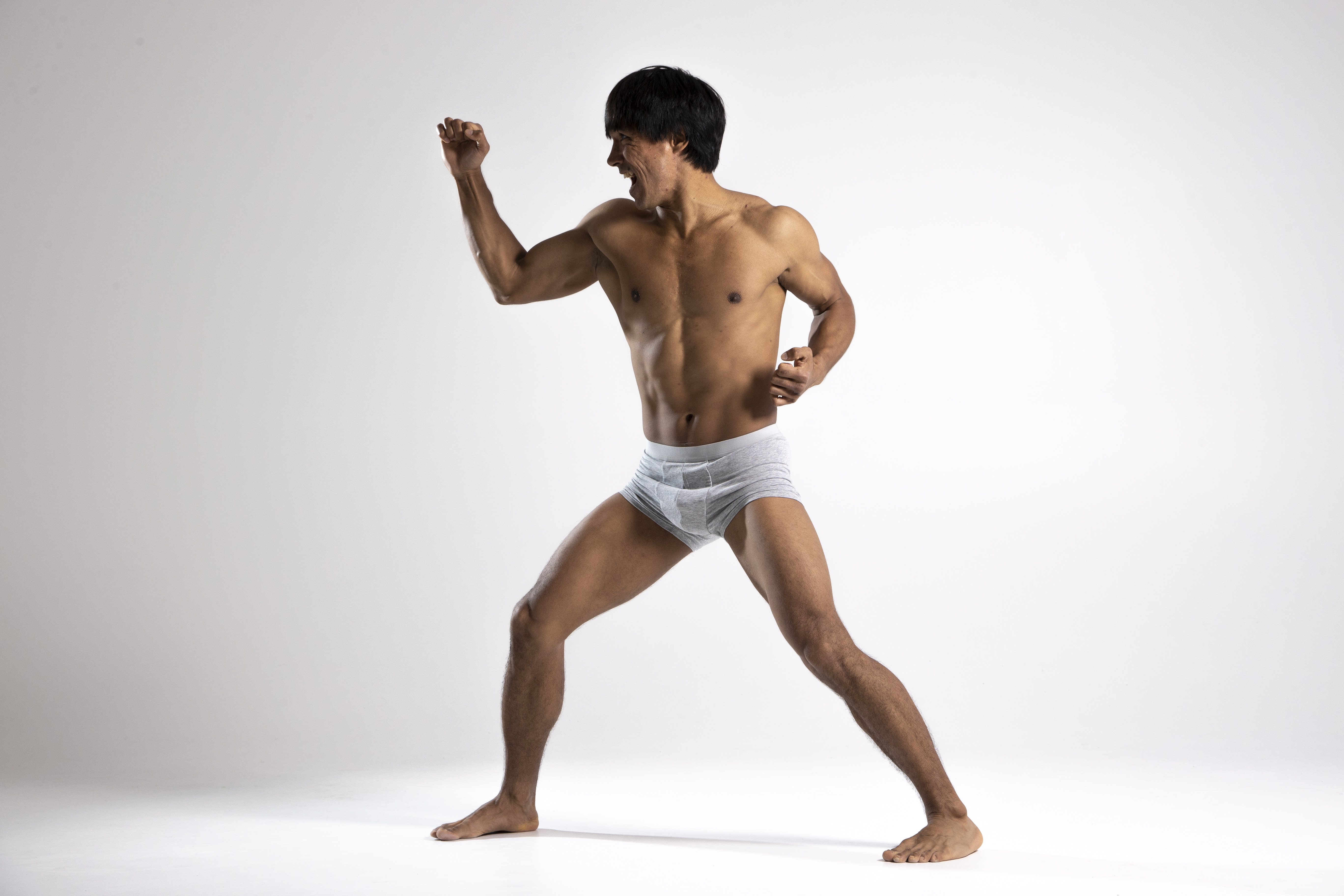 a shirtless asian man wearing grey underwear is in a fighting stance against a white background. he is muscular with dark hair and is barefoot. he has one fist raised in the air and is clenching his other fist as well.