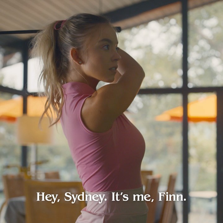 the image shows a young blonde woman, possibly in her late teens or early twenties, from the side profile.  she's wearing a pink sleeveless golf shirt and is in a stylish, modern home with large windows offering a view of a sunlit outdoor area, possibly a golf course or resort.  she's holding a golf club, seemingly preparing to take a swing, and her expression is one of concentration.  text overlayed on the image reads, "hey, sydney. it's me, finn." suggesting a scene from a film or tv show. the overall mood is one of relaxed confidence and leisure.