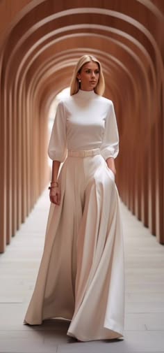a beautiful woman in a white dress stands in a white studio, looking at the camera. she is wearing a white skirt and a white top, with a belt around her waist. she is standing in front of a background of arches. the woman is posed in a relaxed yet elegant manner, highlighting her fashion and beauty.