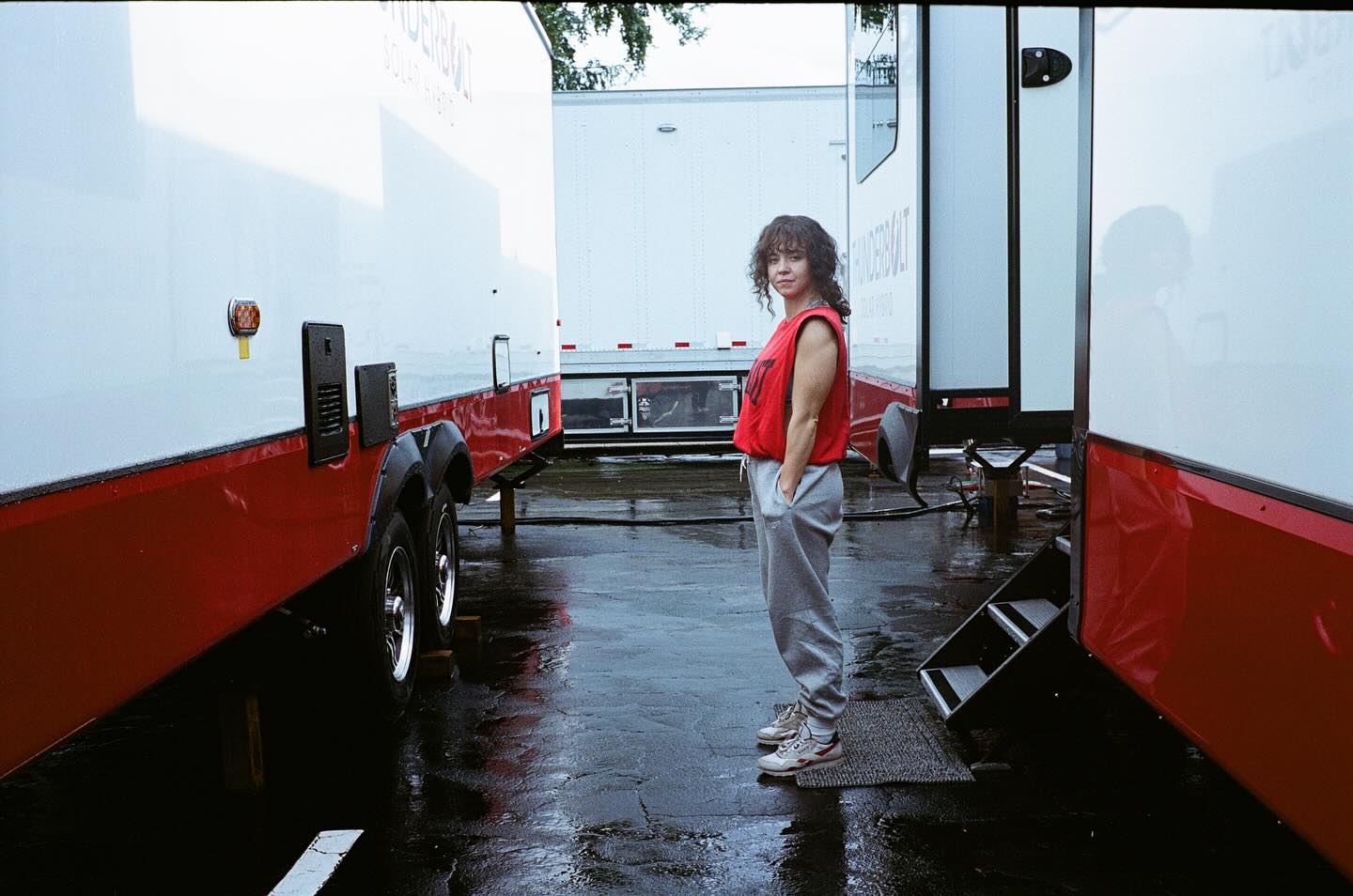 a woman with curly brown hair is standing on a wet, outdoor film set. she is wearing a red tank top and grey sweatpants and casual shoes. in the background are several large red and white trailers typically used for film and movie production. the ground is wet indicating it might have rained.  the overall feel is behindthescenes of a movie production.