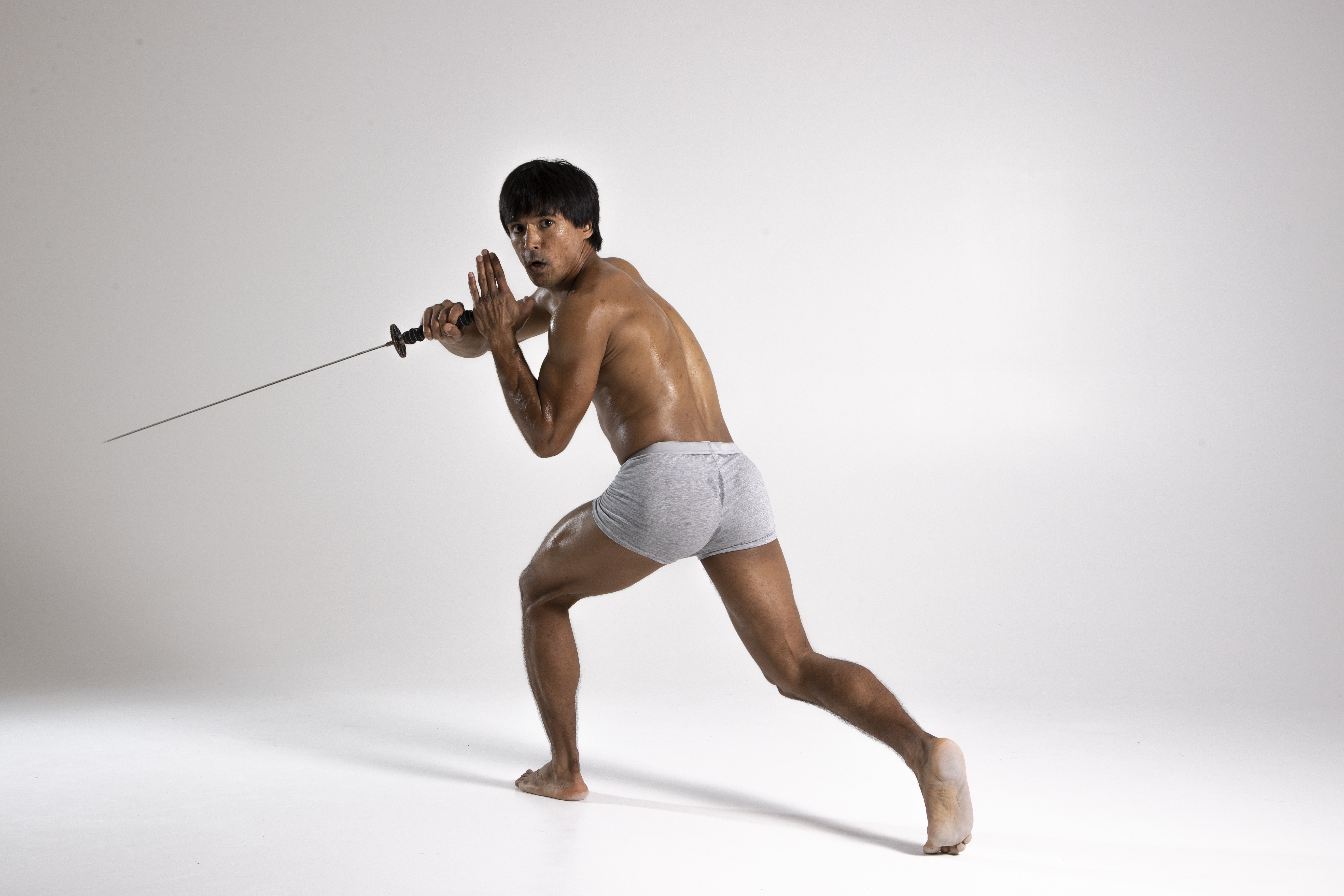 a shirtless man in grey shorts strikes a fighting stance, holding a sword with both hands in a studio shot. he is barefoot on a white background.