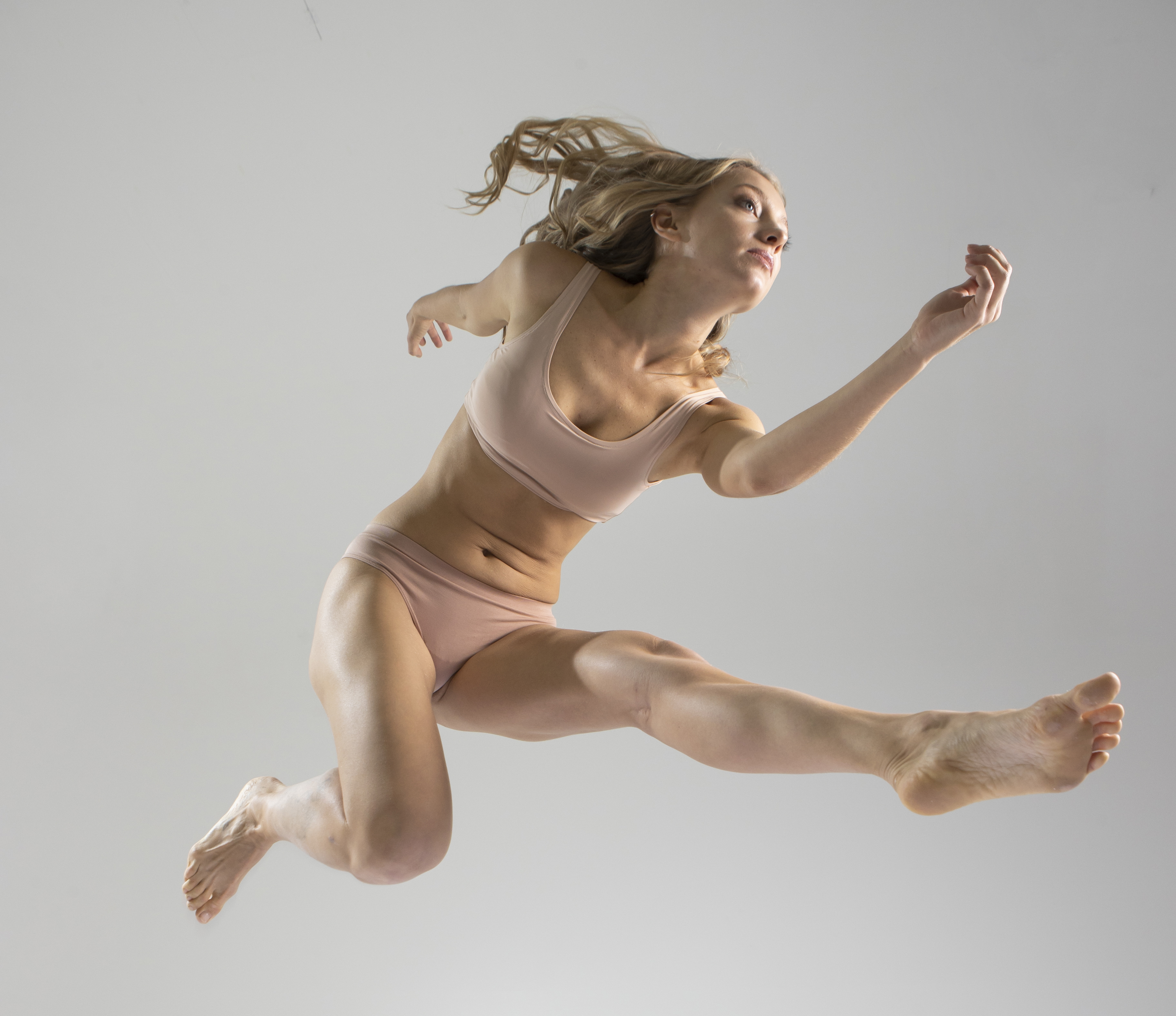 a blonde woman in a nudecolored sports bra and underwear jumps in the air. her hair is in motion, she is barefoot, and she appears athletic and fit. the background is a plain light gray. the overall impression is one of active lifestyle and body positivity.