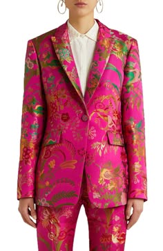 a woman in a pink floral patterned blazer and matching pants stands with a slight smile on her face. she is wearing a white shirt beneath the jacket. the floral pattern on the blazer and pants is intricate and detailed. the woman is wearing a pair of small hoop earrings.