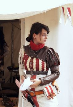 a woman dressed in a medieval or renaissancestyle costume is standing in or near a tent. she appears to be looking down and to the right, with a thoughtful expression on her face. she is holding a white cloth in her left hand and the straps of a satchel in her right hand.  the costume features a red and white striped bodice with brown leather accents, a red scarf, and a white skirt. the background suggests she is at an outdoor event, possibly a renaissance fair, and the lighting suggests it is daytime with natural light illuminating the scene.
