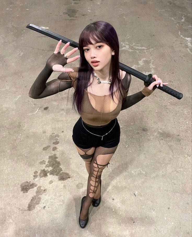 a young asian woman wearing black fishnet stockings, a black crop top, black short shorts, and black pumps poses with a katana sword. the woman looks directly at the camera, her dark hair falls over her shoulders. she is posing in a studio setting, with a concrete floor in the background.