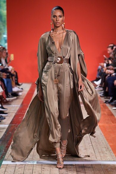 a fashion model walks down a runway wearing a stylish beige silk jumpsuit with a matching long over coat. the jumpsuit is cinched at the waist with a brown leather belt adorned with a large gold buckle. the model wears delicate gold jewelry including a long necklace, earrings, and a few stacked bracelets. her hair is styled in a sleek updo. the model's highheeled, embellished sandals and minimal makeup complete the look, giving a overall elegant and glamorous feel. the background features a red backdrop and other audience members. the setting appears to be a fashion show or presentation of a new collection.