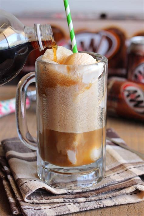 a closeup shot of a root beer float being made. a glass mug is filled with ice cream and root beer. a bottle of root beer is being poured into the mug. a green and white straw is in the mug. the mug is sitting on a folded plaid cloth napkin. the background is blurry, with some root beer cans out of focus.
