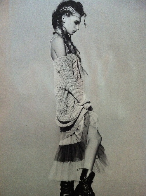 a black and white photo of a female model with her back to the camera. she is wearing a white sweater, a tutu skirt, and black boots. her hair is in braids. the photo is taken in a studio setting.