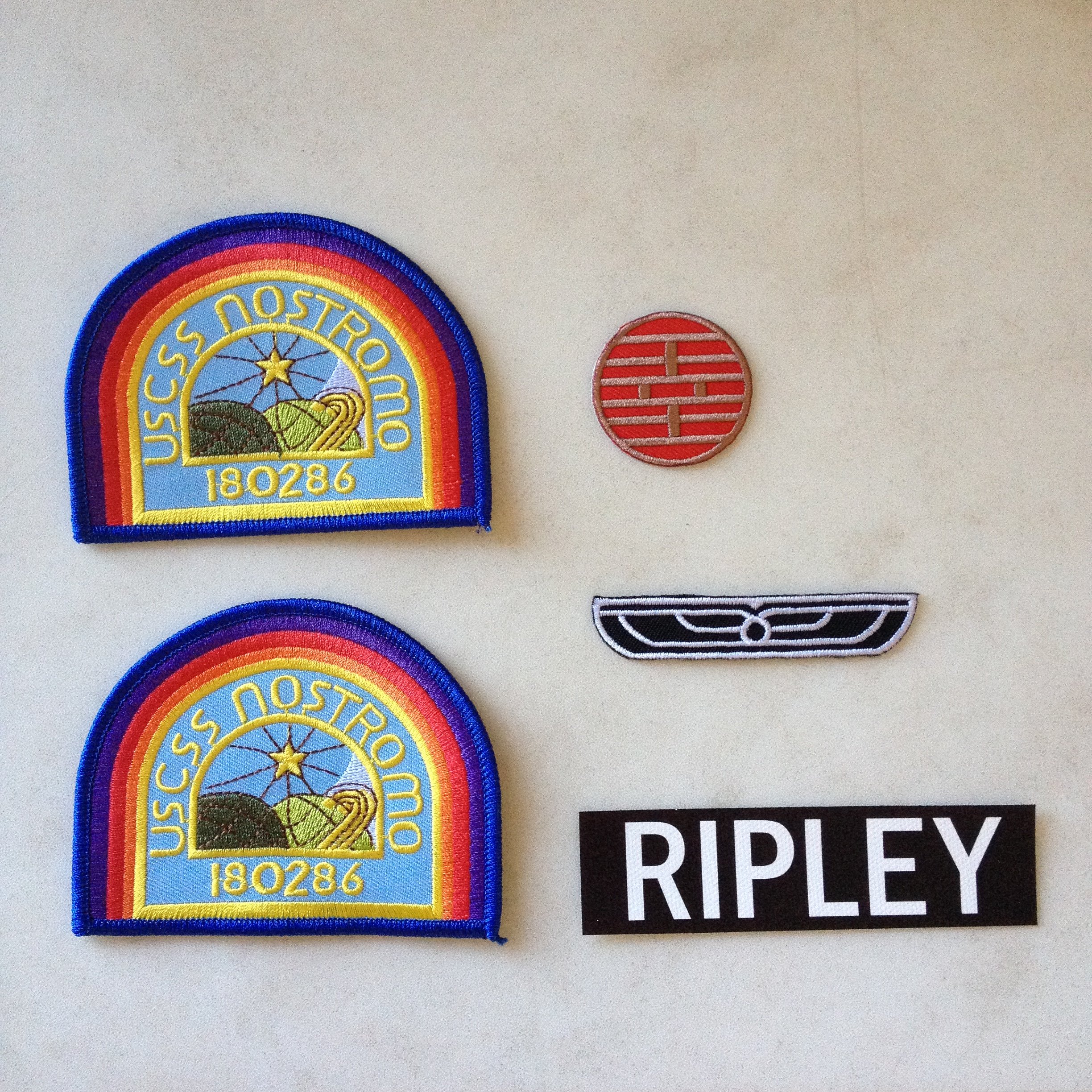 this is a picture of 5 patches from the movie alien, they all have the name "nostromo" on them along with the uscs logo, except one which is a simple black patch with the word "ripley" on it in white letters. the other 4 patches have the same design but with different color combinations, and have the number 180286 on them. the patches seem to be sewn onto the image's background, which looks like a piece of fabric.