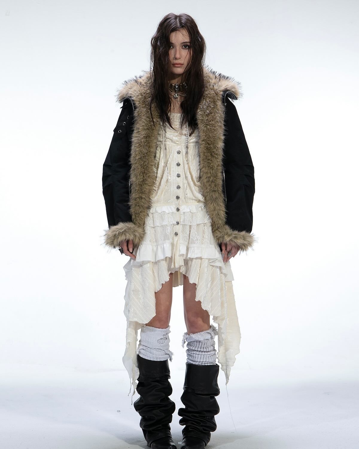 a full shot of a female model on a white background, wearing a black parka with a furtrimmed hood, a white ruffled dress, white kneehigh socks, and black slouch boots.  the overall style is a mix of grunge, bohemian, and edgy. the model has long dark hair and a neutral expression. the image appears to be from a fashion show or photoshoot.