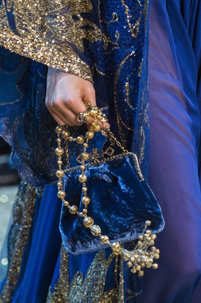 loseup shot of a woman's hand holding a luxurious blue velvet handbag with an ornate gold chain and embellishments. the handbag is a rich, deep blue velvet, and the chain is decorated with golden beads and intricate detailing.  the woman is wearing a matching long blue gown heavily embellished with gold sequins and embroidery. the overall impression is one of luxury, elegance, and high fashion. the image focuses on the texture and detail of the handbag and the gown's fabric.