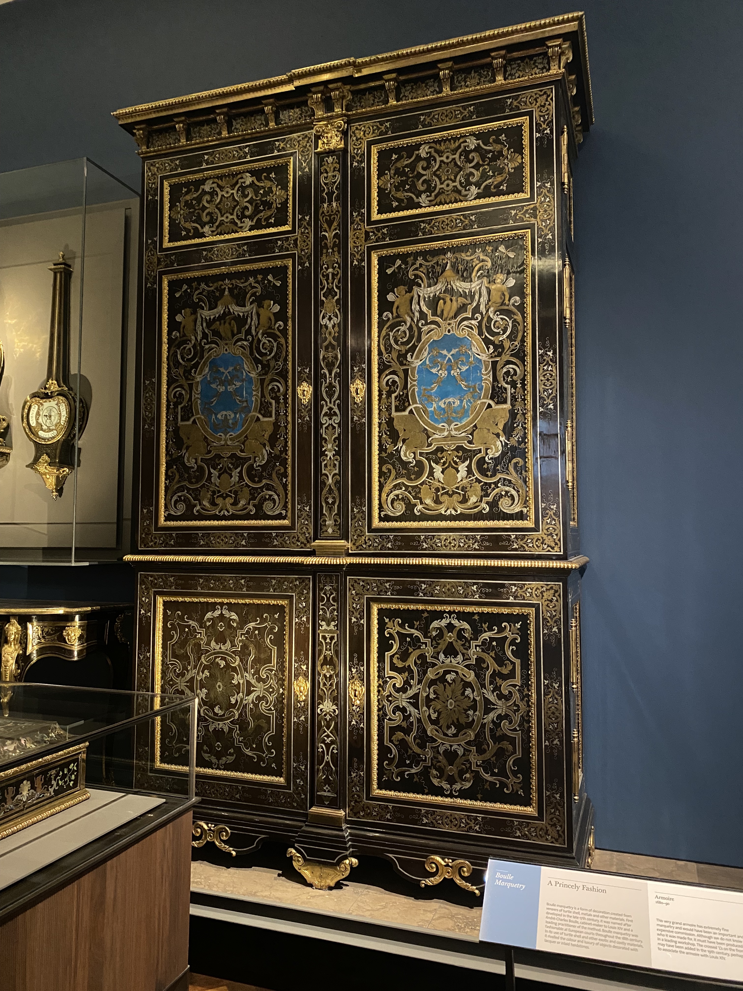 a large antique cabinet with ornate gold trim and a blue shield emblem is displayed in a museum setting. the cabinet has multiple doors and intricate designs throughout.