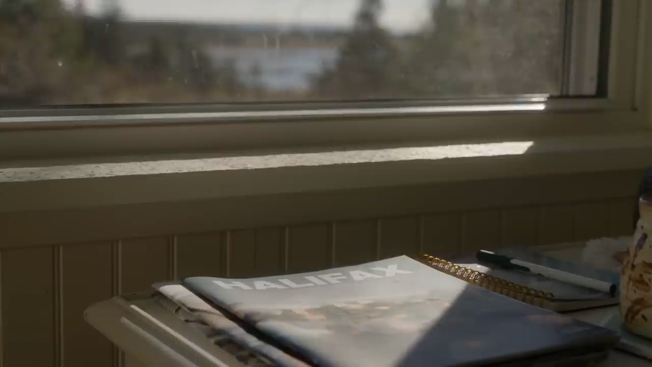 a sunlit interior with a magazine and notebook lying on a table in front of a window. there's a view of trees and water outside the window.