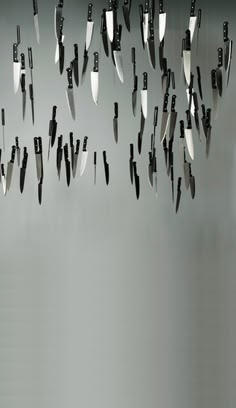 a white wall with hundreds of knives of varying sizes hanging, suspended, and pointed downwards. the wall appears to be a grey color.  a stark and abstract art installation.