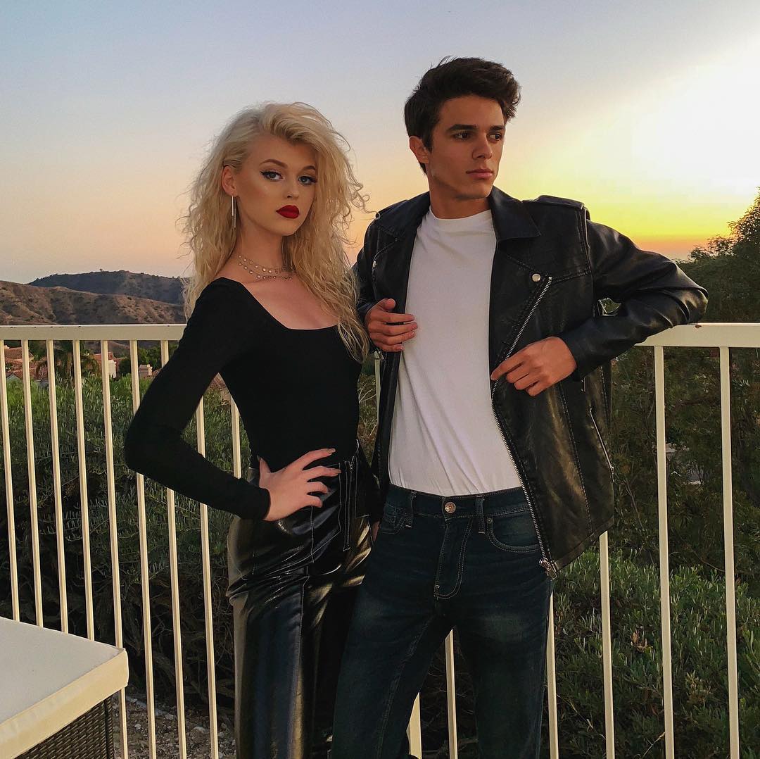 a woman with blonde curly hair and a man with short dark hair stand in front of a railing at sunset. the woman is wearing a long sleeve black top and black leather pants. the man is wearing a white tshirt and a black leather jacket with dark jeans.