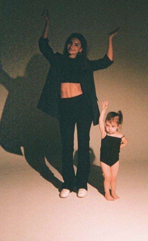 a full shot of a woman and a young girl posing in a studio. the woman is wearing a black blazer, crop top, and black pants. the girl is wearing a black leotard. they are both smiling and appear to be enjoying themselves. the background is a light beige color. the image has a vintage or filmlike quality.
