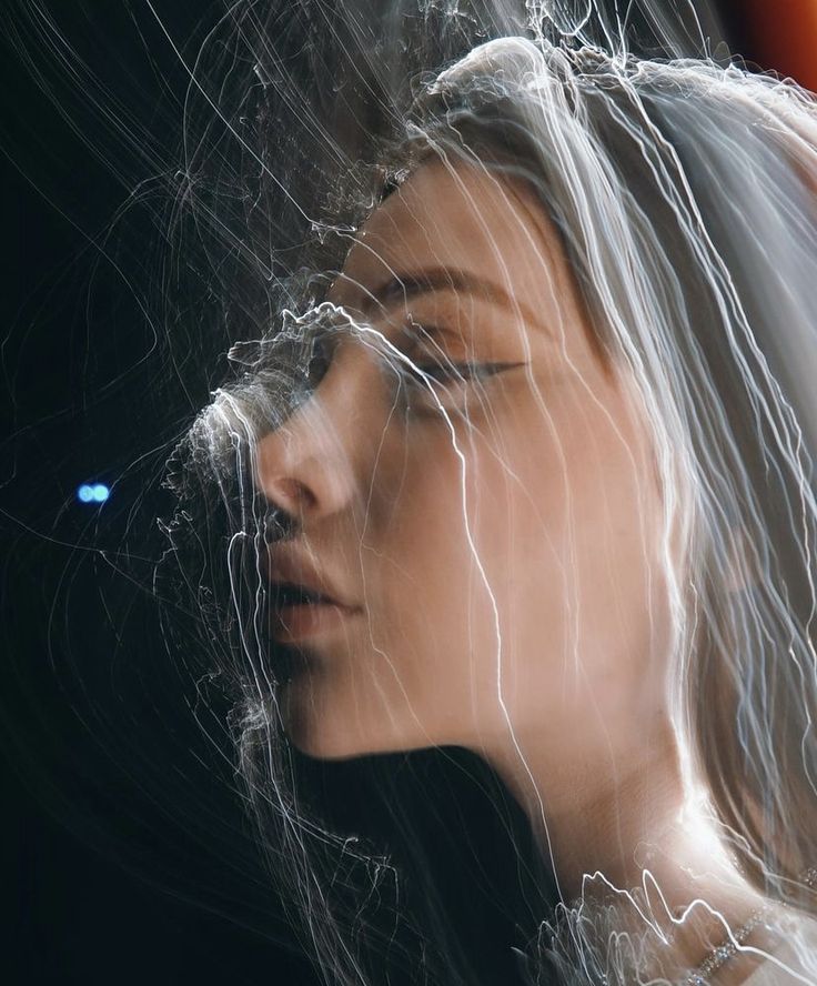 the image is a stylized portrait of a woman's face.  it's a long exposure photograph featuring light painting techniques where streaks of white light appear to flow around and through her face and hair, creating an ethereal and somewhat abstract effect.  her eyes are closed, and her expression is serene. the background is dark, drawing attention to the glowing light trails that intertwine with her features. the overall mood is mysterious and dreamy.