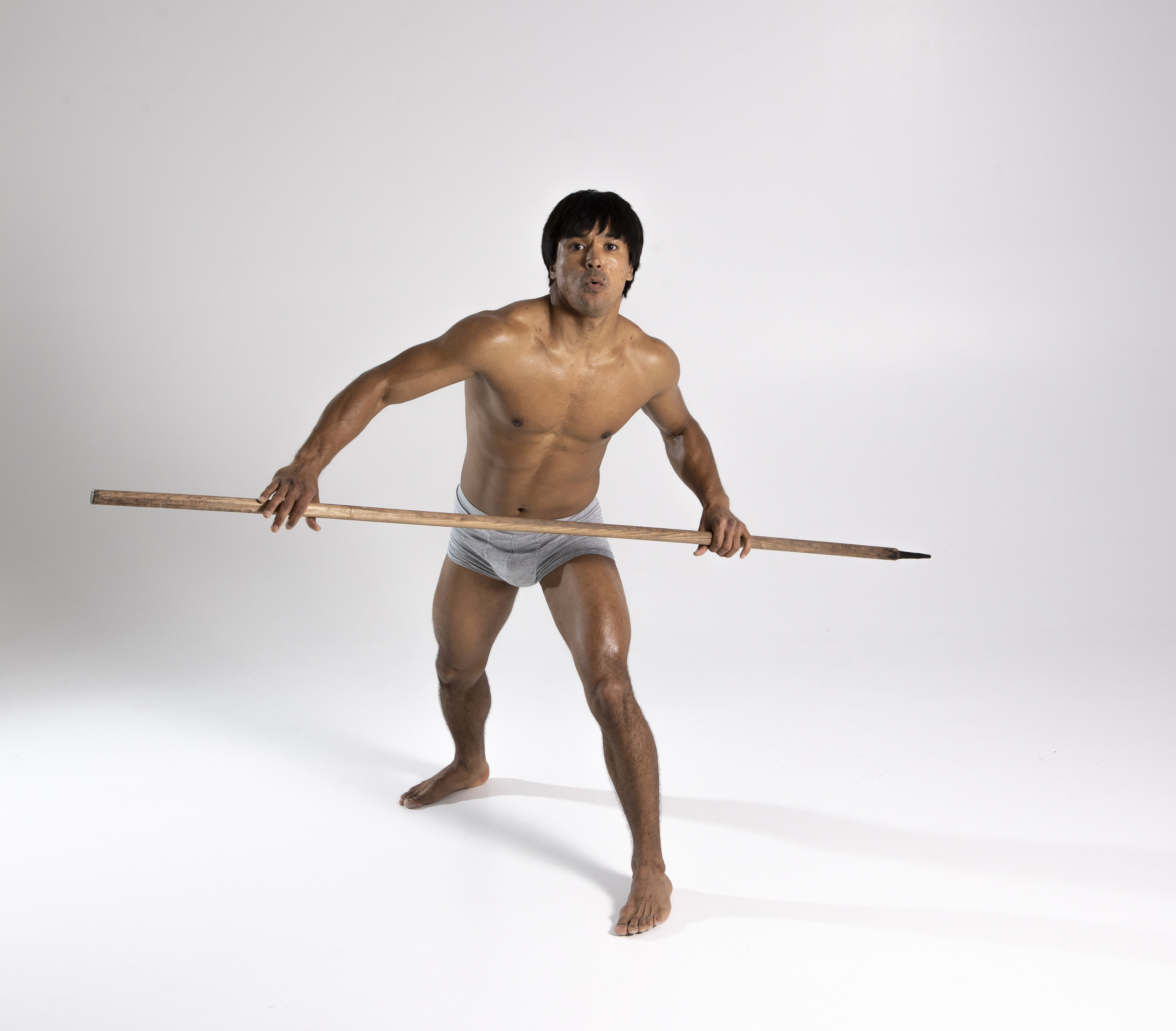 a shirtless muscular man wearing underwear is holding a spear while looking at the camera. he is standing in a defensive pose with his bare feet on a white background. he has brown hair and is posing indoors in a studio.