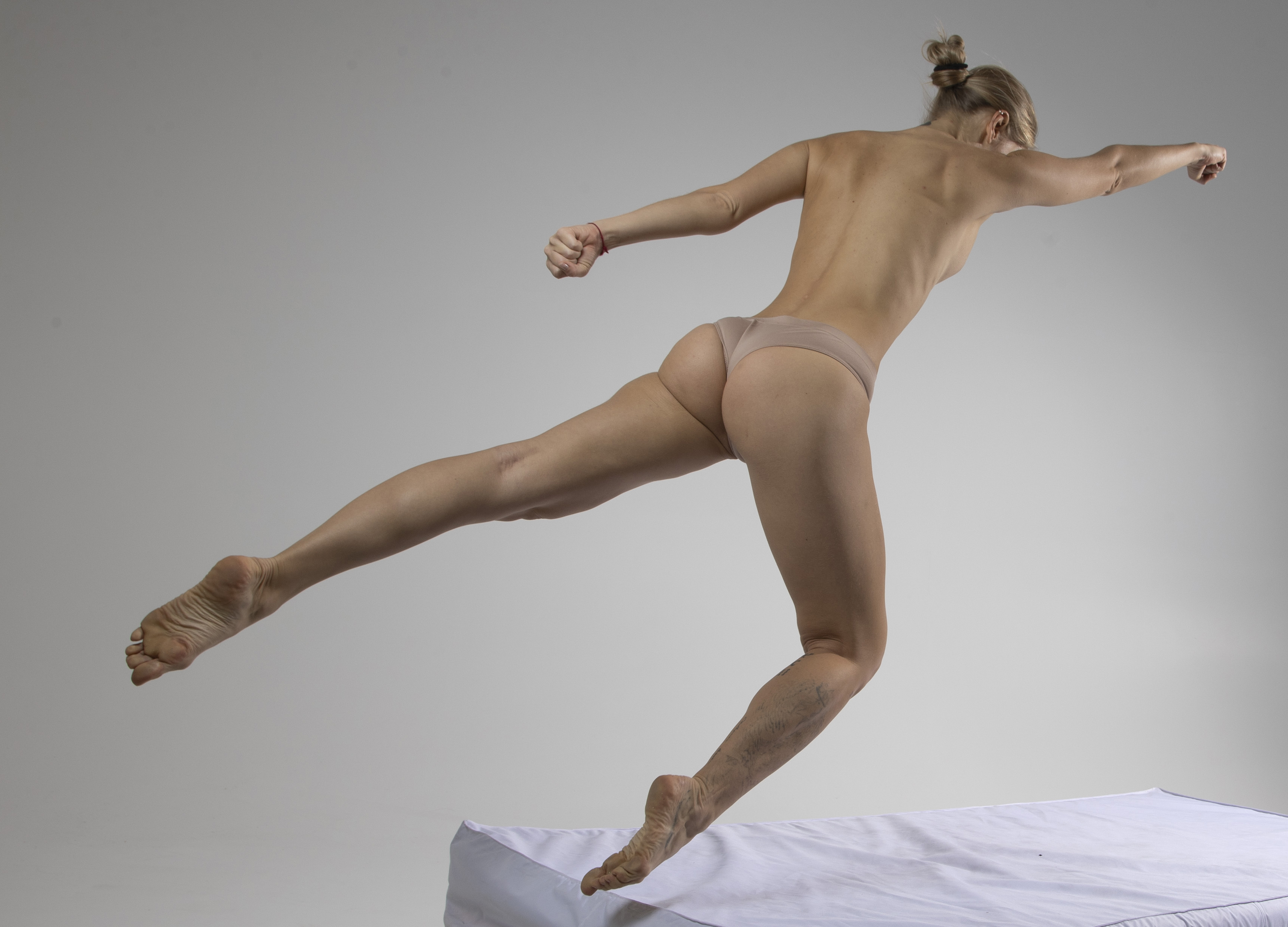 a full shot of a woman with a bun hairstyle, posing nude except for beige underwear, in an athletic jumping pose against a white background. she has a toned body and is viewed from the back.