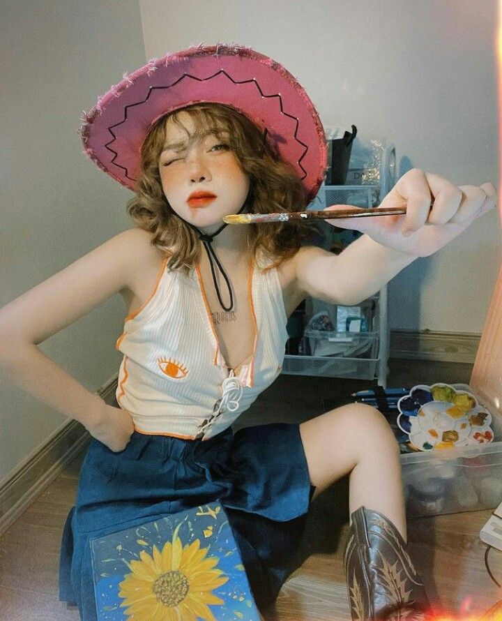 a young woman wearing a cowboy hat and a white tank top and blue shorts is holding a paintbrush and smiling. she is sitting on the floor, and a painting is leaning against her legs.  she is wearing a pink cowboy hat. the painting shows a sunflower.  she looks happy and carefree.  she seems to be an artist.