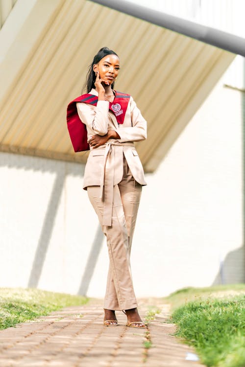 a full shot of a stylish, young black woman in a beige pantsuit and maroon graduation stole. she stands confidently on a brick pathway outdoors, smiling slightly. the backdrop features a modern white building.  the overall impression is one of accomplishment and elegance.