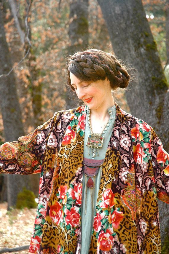 a woman is wearing a long, colorful floral kimono with a leopard print lining. she is standing in a forest and looking down. she is smiling and wearing a large, ornate necklace.