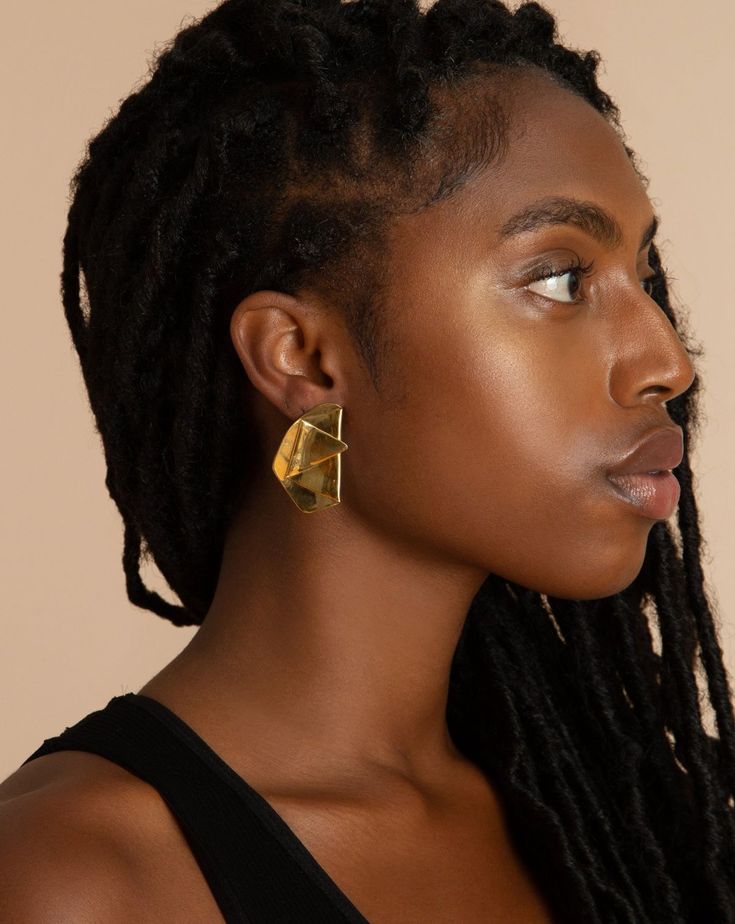 a profile shot of a black woman wearing a large gold geometric earring. she has dreadlocks and is wearing a black top. the background is a tan color. her skin is glowing.