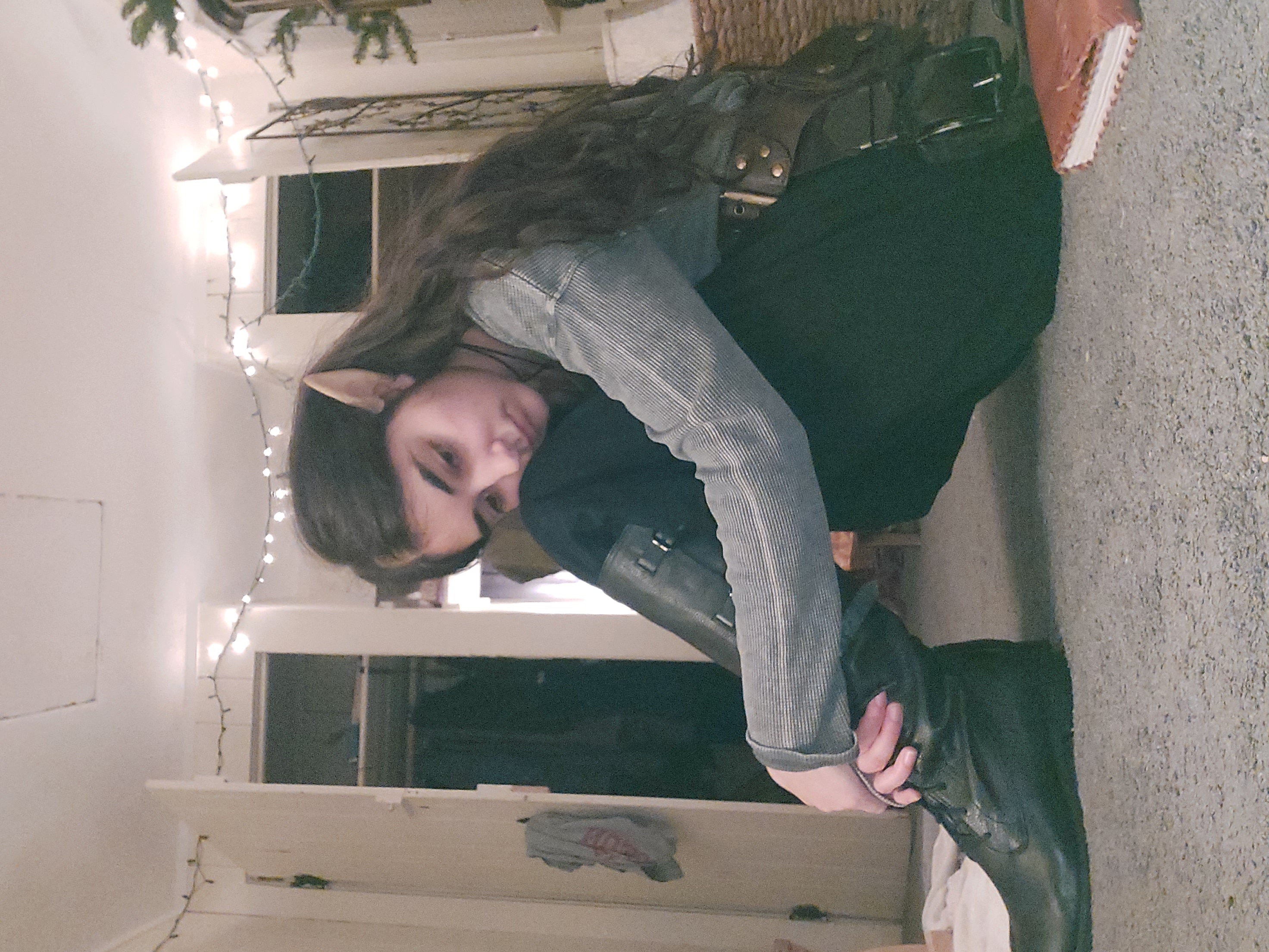 a young woman, possibly a teenager, dressed in an elf costume is sitting on the floor.  she's wearing elf ears, a grey sweater, black pants, and tall black boots.  she appears pensive or sad, with her head bowed.  the setting is an indoor room with string lights and a visible closet in the background.  a journal or notebook lies on the floor near her. the overall mood is somewhat melancholic and evocative of fantasy or roleplaying.