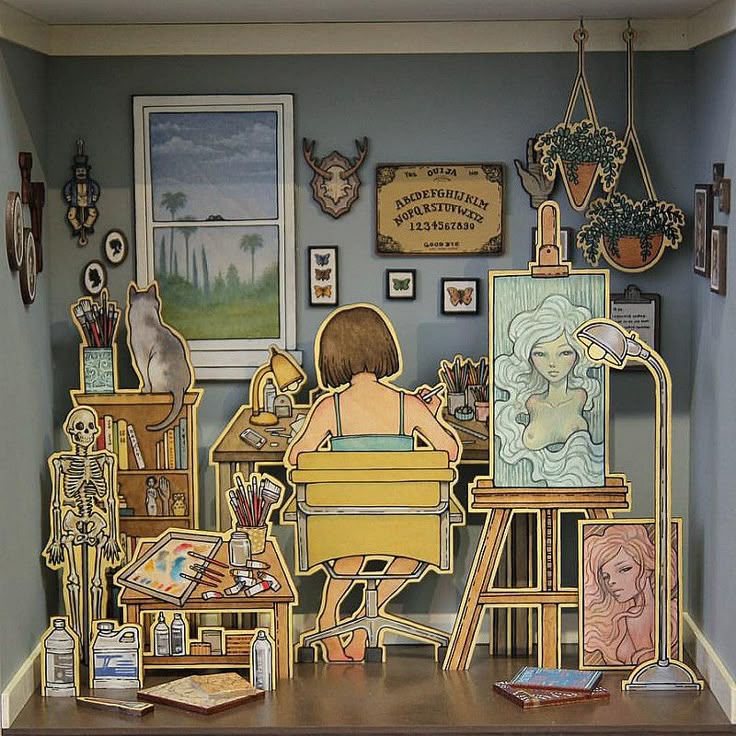 a dollhousestyle image depicts an artist's studio created with paper cutout illustrations, featuring a person painting at an easel, a cat sitting on a bookshelf, and various artistic elements such as paintbrushes, paintings, and a skeleton model. the room is decorated with cutouts of artwork, a window, hanging plants, and a ouija board.