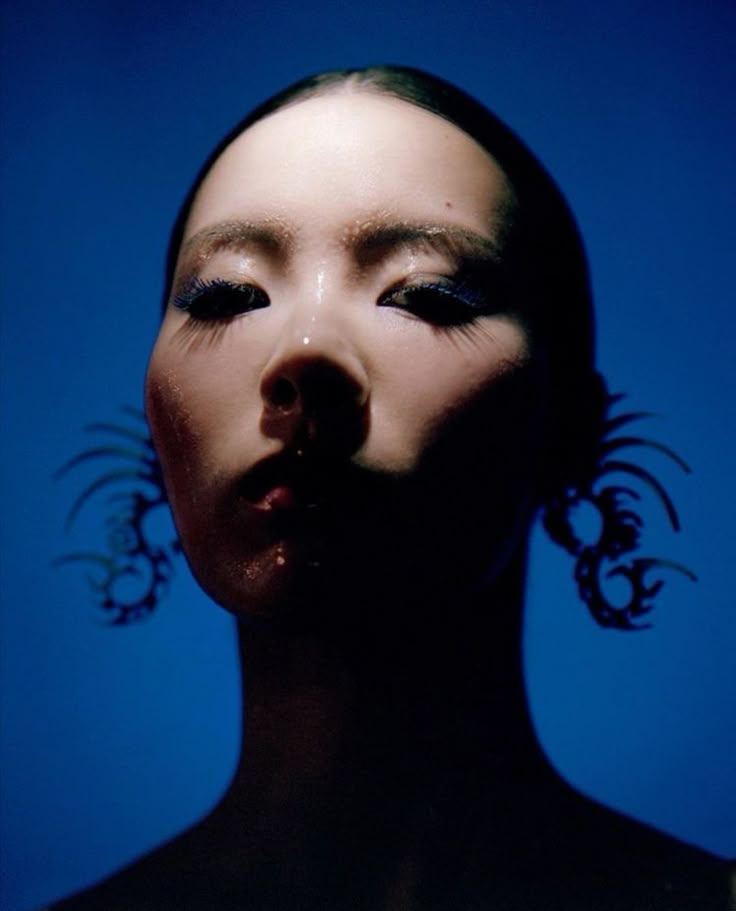 a portrait of an asian woman with slickedback hair against a blue background. she has a glossy, wet look with glitter on her face and eyebrows. she is wearing ornate earrings and makeup that includes blue eyeliner. the lighting is low key, creating strong shadows.