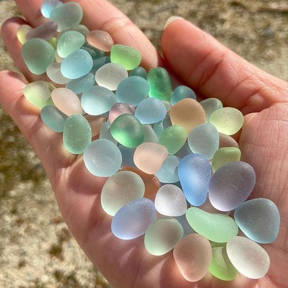 a closeup of a hand holding a collection of smooth, colorful sea glass. the glass is a variety of shades of blue, green, and pink. the hand is holding the sea glass in the palm of their hand, and it is resting on a sandy beach. the sea glass is a beautiful reminder of the ocean and its treasures.