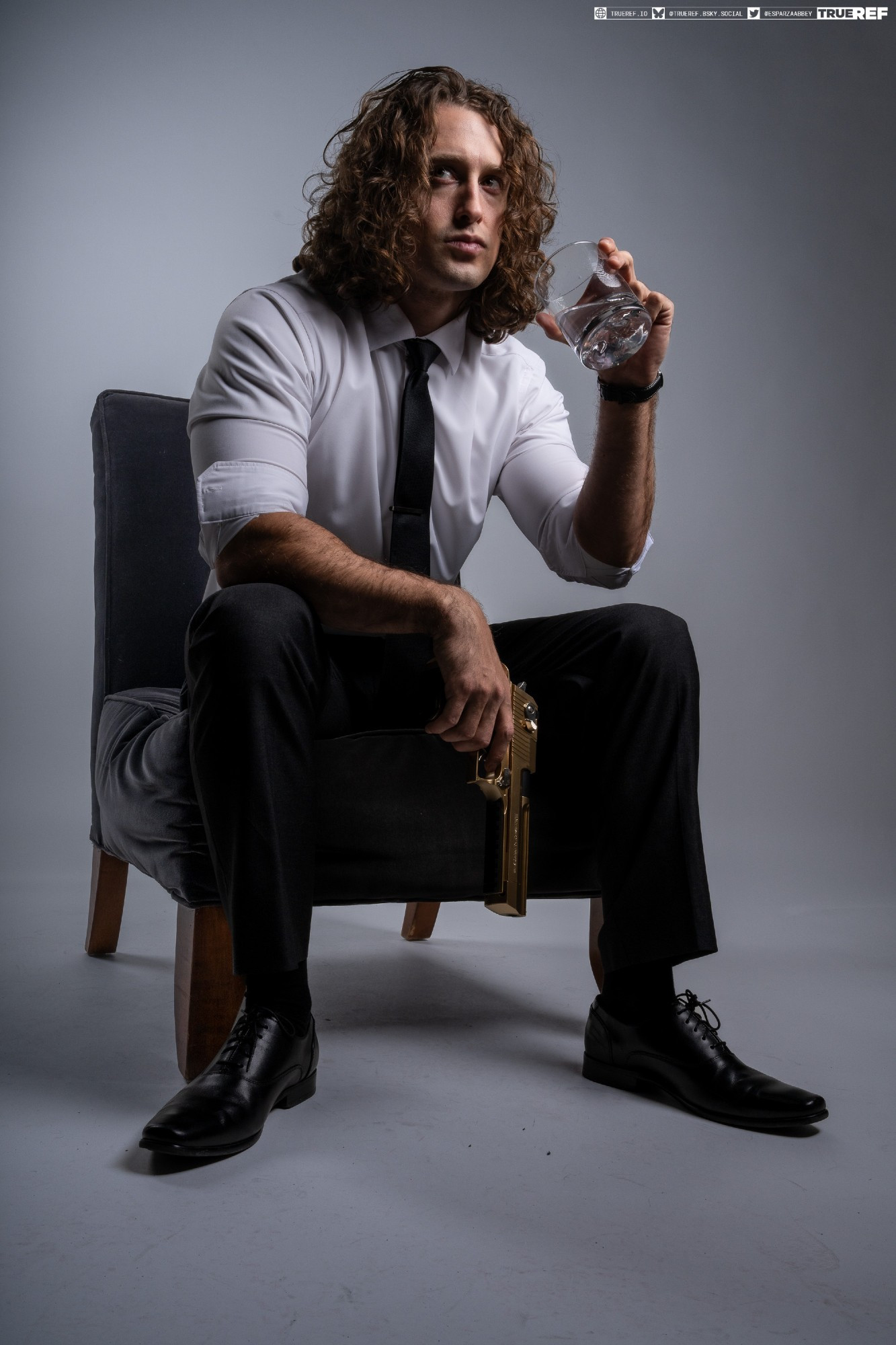 the image shows a young man with long curly brown hair, dressed in a sharp black suit and white shirt with a tie, sitting thoughtfully in a dark grey armchair. he holds a goldcolored pistol in his lap and is holding a glass of water to his lips. his expression is serious and pensive, creating a mysterious and somewhat brooding atmosphere. the background is a simple, even grey, focusing attention on the subject. the overall style and pose suggest a character from a crime drama or thriller.