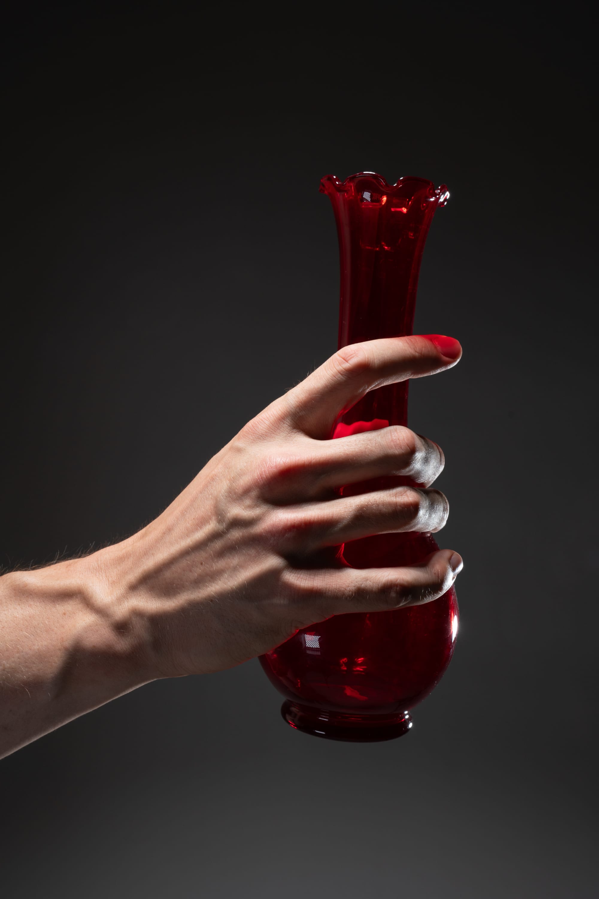 male hand is dramatically lit from the side, holding a red glass vase with a flared lip. the vase is empty, and the hand is lightly stained with red, suggesting recent use. the background is a dark gray, emphasizing the contrast of light and shadow.