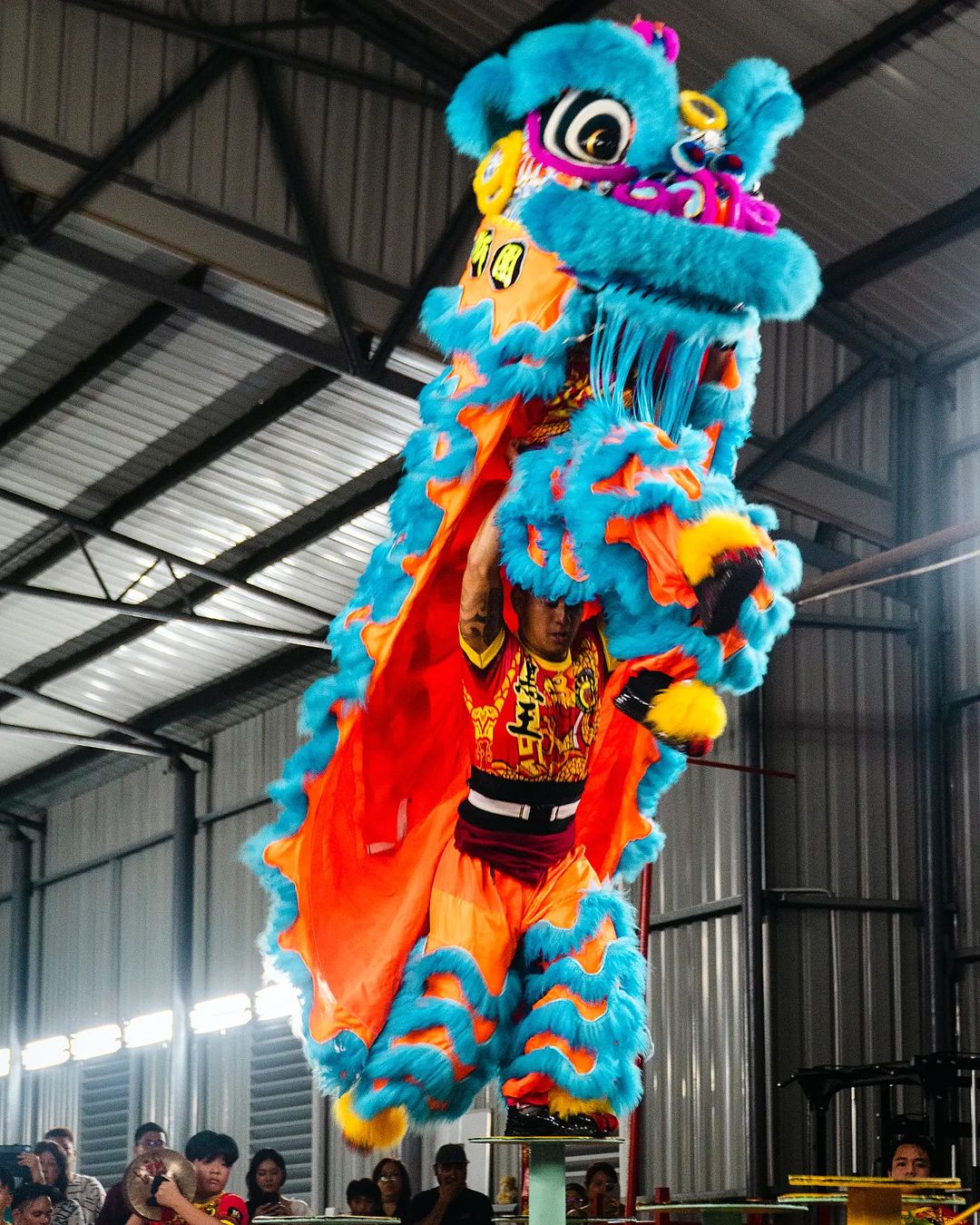 a single lion dancer stands atop a pole midperformance.  the vibrant orange and blue costume is highlighted by the artificial lights of the large industrial building the performance is taking place in. the crowd below watches expectantly.