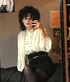 a woman dressed in a black skirt and white shirt stands in front of a mirror with a serious expression. she's wearing a silver belt with a black necklace. her black hair is styled with a side part, and she's wearing a black dress. she looks up at the camera. her reflection in the mirror is visible behind her.