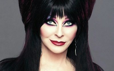 a woman with dark black hair and heavy dark eye makeup is looking at the camera. she is wearing a costume with black hair, dark eyeliner, black lipstick, and a black dress.