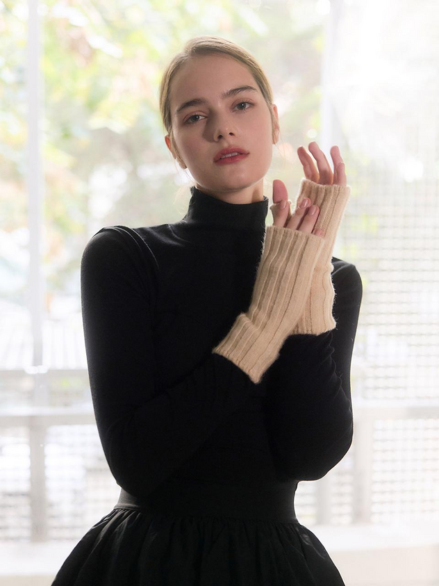 a woman is posing in a studio wearing a black turtleneck and a black skirt. she is wearing beige fingerless gloves and has her hands up in front of her. there is a window behind her with blurred greenery visible through the glass.