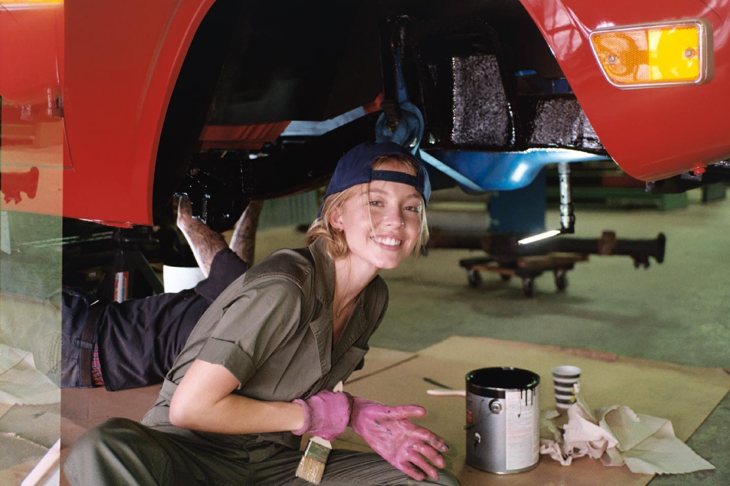 a young woman, a mechanic, smiles at the camera while working under a raised red car in a garage. she's wearing work overalls, gloves, and a cap, and is holding a paintbrush and what appears to be a can of paint. another person's legs and lower body are visible under the car, suggesting teamwork. the scene depicts a classic car undergoing repair and painting. the overall mood is positive and professional.
