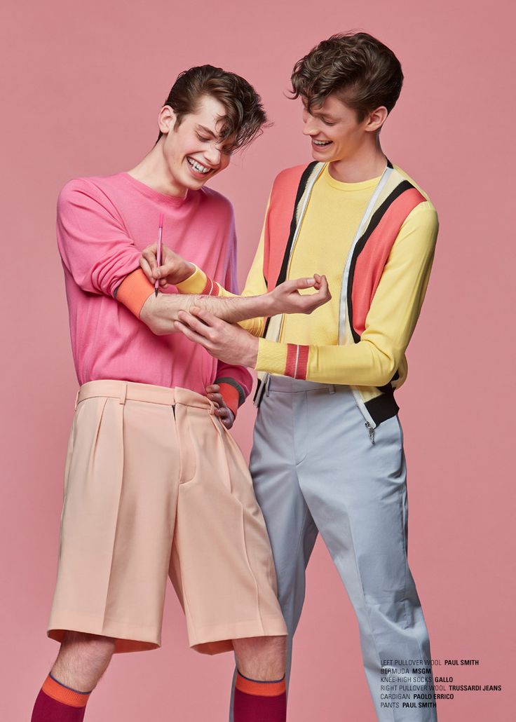 the image shows two young men in colorful clothing, posing for a photo shoot. the man on the left is wearing a pink sweater and pink shorts with socks. the man on the right is wearing a yellow sweater with a red stripe over a white tshirt and blue pants. the man on the left is drawing on the arm of the man on the right, as the man on the right is looking at him and smiling. the backdrop is a soft pink. both models are smiling and looking at each other, creating a playful, youthful energy.