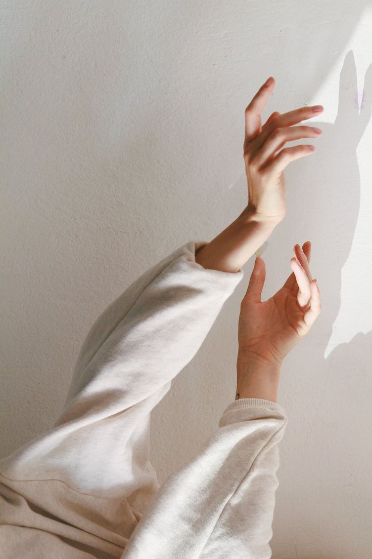 a person's hand is reaching up towards the light. the other hand is visible at the bottom of the frame.  the person is wearing a white sweater and the sunlight is casting a shadow on the wall.  the light is bright and the background is simple.  the overall feeling is one of peace and serenity.