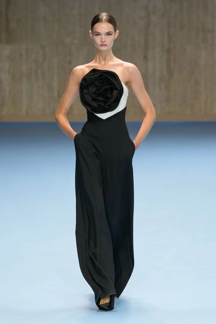 a female model walks the runway wearing a black strapless jumpsuit with a white and black rose on the bodice. her hair is pulled back and her makeup is minimal. she is accessorizing with black heels. the runway is a light blue color.