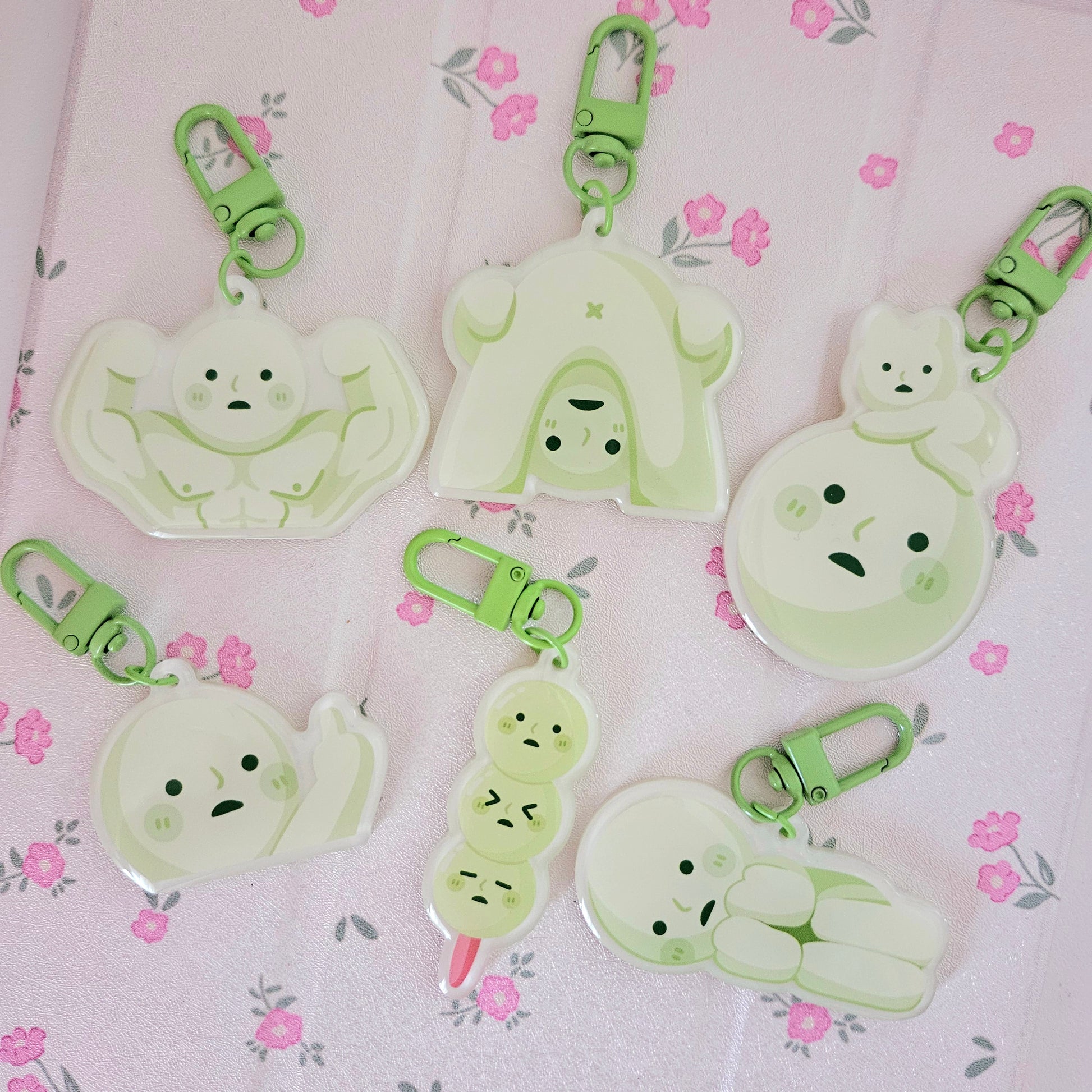 a flatlay photograph showcasing a collection of six adorable, pastel green keychains.  each keychain features a different whimsical character design, ranging from a muscular ghost to a mochi character carrying a smaller mochi.  the keychains are made of translucent acrylic or plastic and have light green metal clasps. the keychains are arranged on a light pink fabric background with a floral pattern. the overall aesthetic is cute, kawaii, and whimsical.