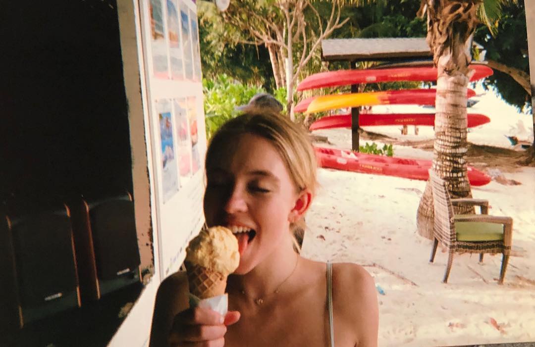 a young woman with blonde hair is happily eating an ice cream cone on a tropical beach.  she's smiling and appears to be enjoying her treat. in the background are colorful kayaks stored on a rack under a palm tree.  the overall setting suggests a relaxing and fun vacation.  the image has a slightly faded, vintage look, possibly from a photograph.