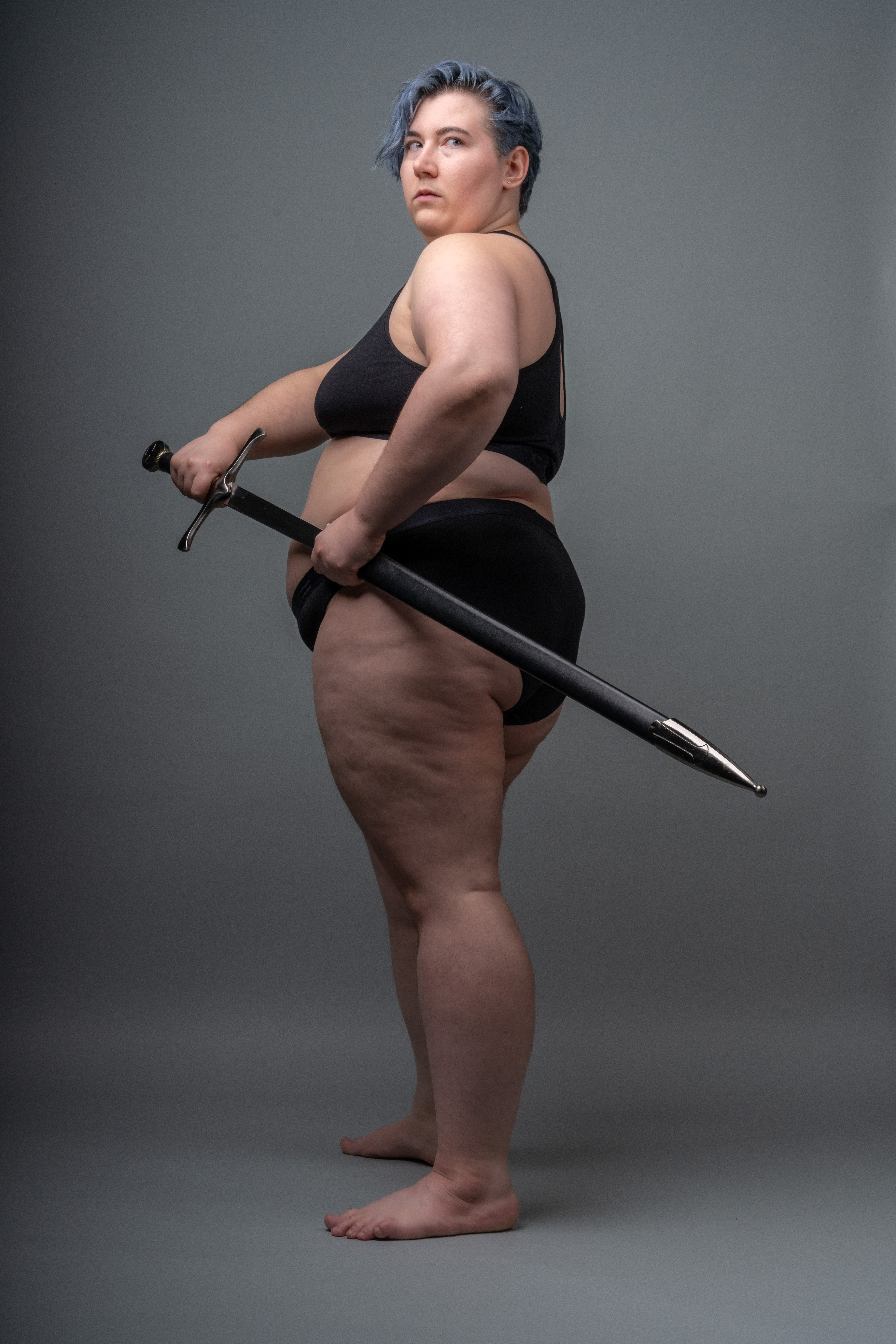 a studio shot of an overweight woman with short blue hair, wearing a black bra and underwear, standing barefoot while holding a sword and looking over her shoulder. she is fairskinned and the background is a solid gray. she is holding the sword in a sheath.