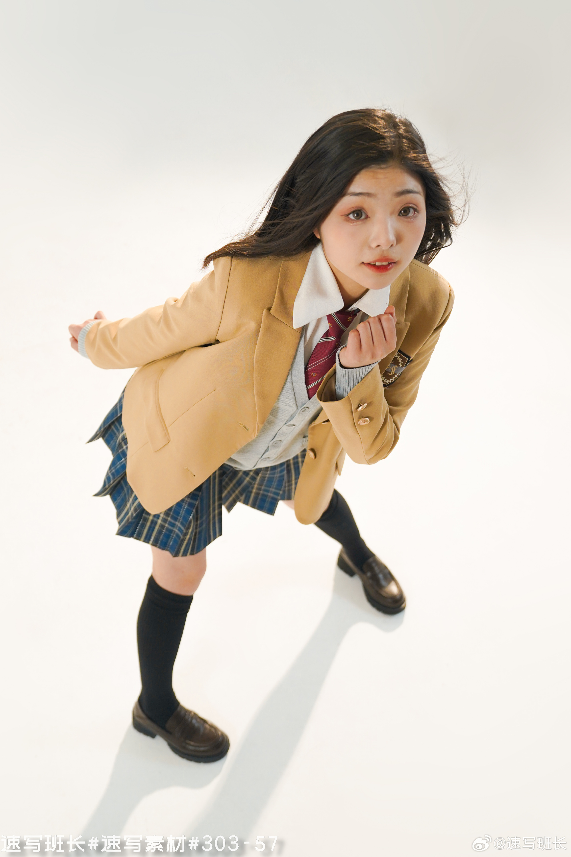 a young asian woman dressed in a school uniform is striking a playful pose against a white background in a studio setting. she is looking directly at the camera with a curious expression. the uniform consists of a tan blazer, a pleated blue skirt, a white shirt, a red tie, black kneehigh socks, and black leather shoes. the lighting is bright and artificial, illuminating her from above, resulting in a highangle perspective.
