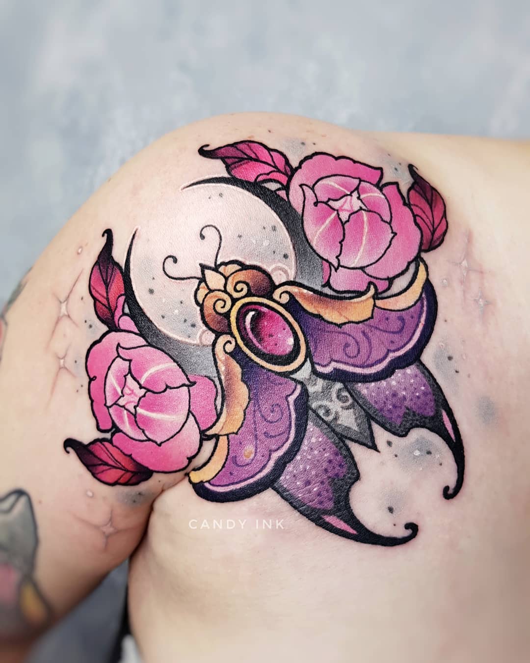 a neo traditional style tattoo of a moth on a shoulder. the moth is purple with gold accents and has a pink jewel in its center. there are pink flowers and a crescent moon behind the moth. there are also some white stars around the tattoo.